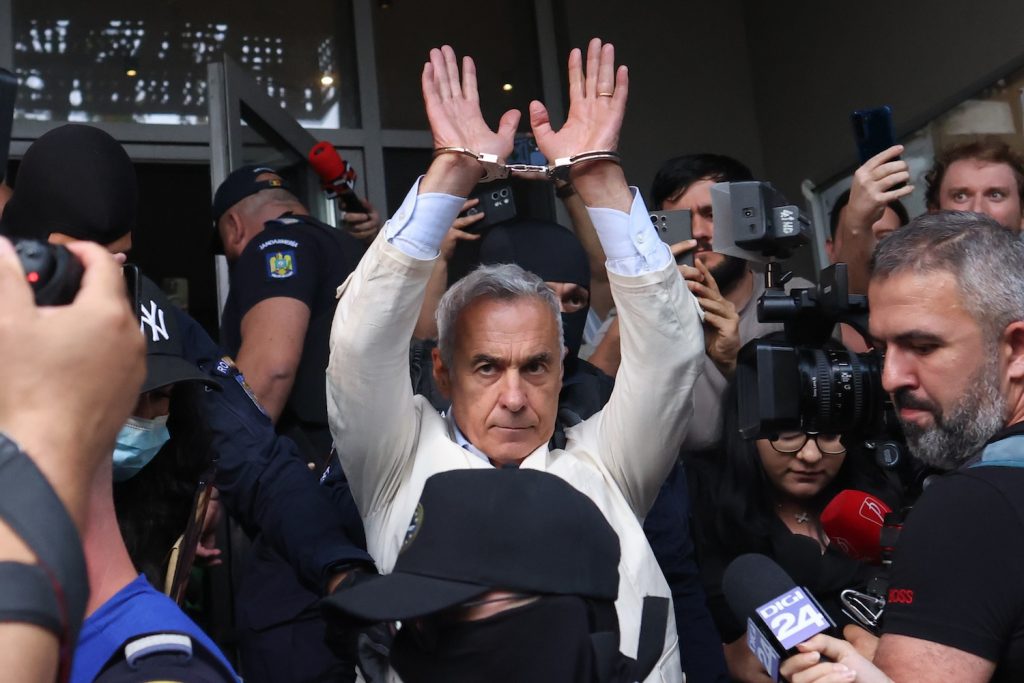 Călin Georgescu, cu mâinile încătușate ridicate, înconjurat de jurnaliști și polițiști