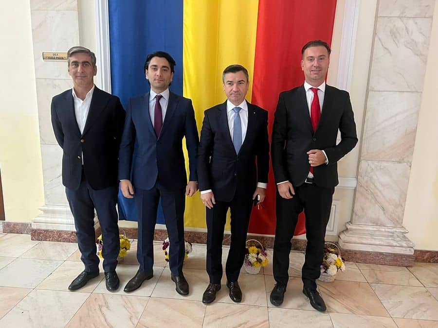 Patru bărbați în costume, în picioare în fața drapelului României, într-o sală cu pereți din marmură și coșuri cu flori pe podea