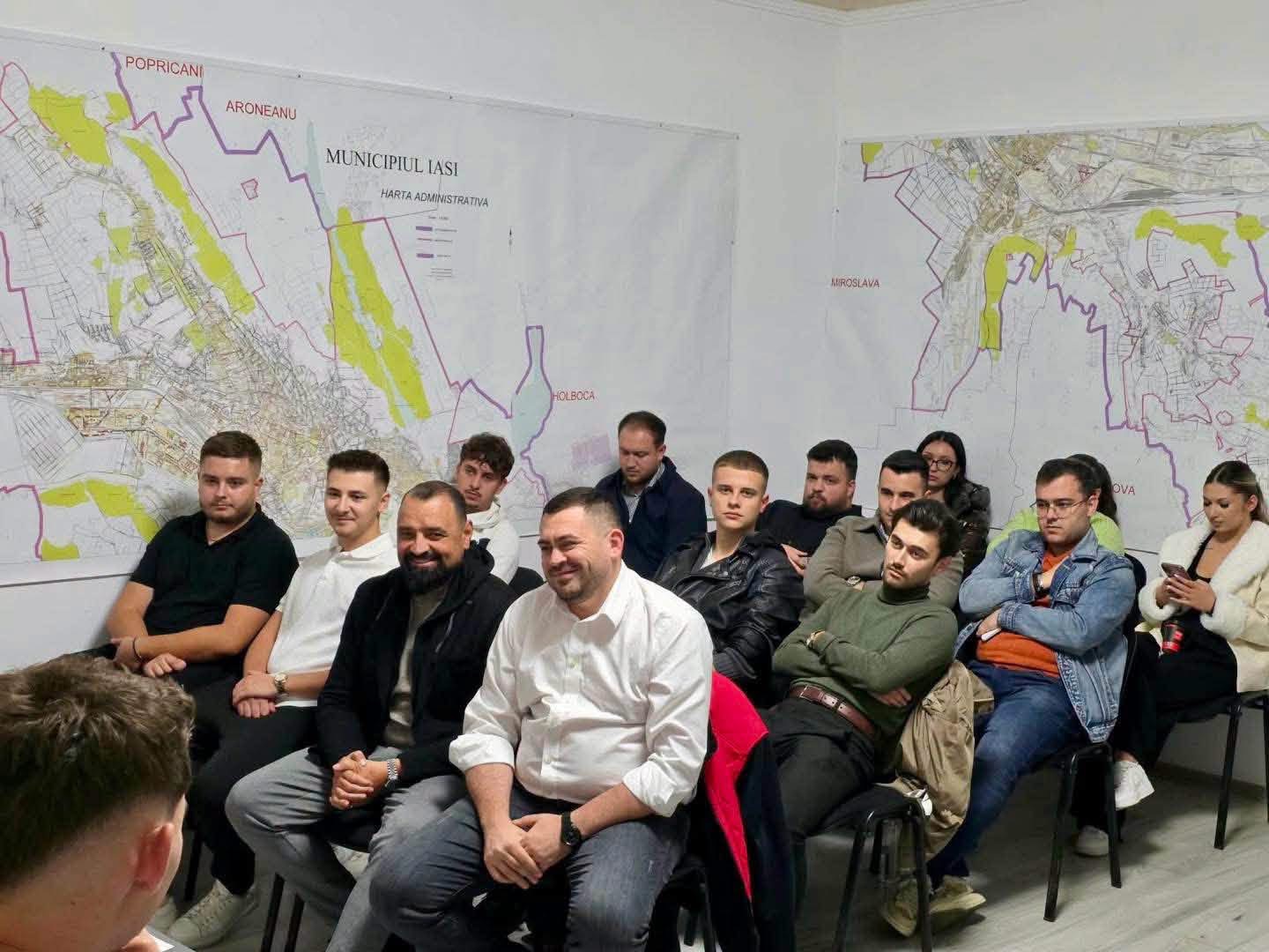 Oameni așezați pe scaune într-o sală cu hărți administrative ale Iașului pe pereți