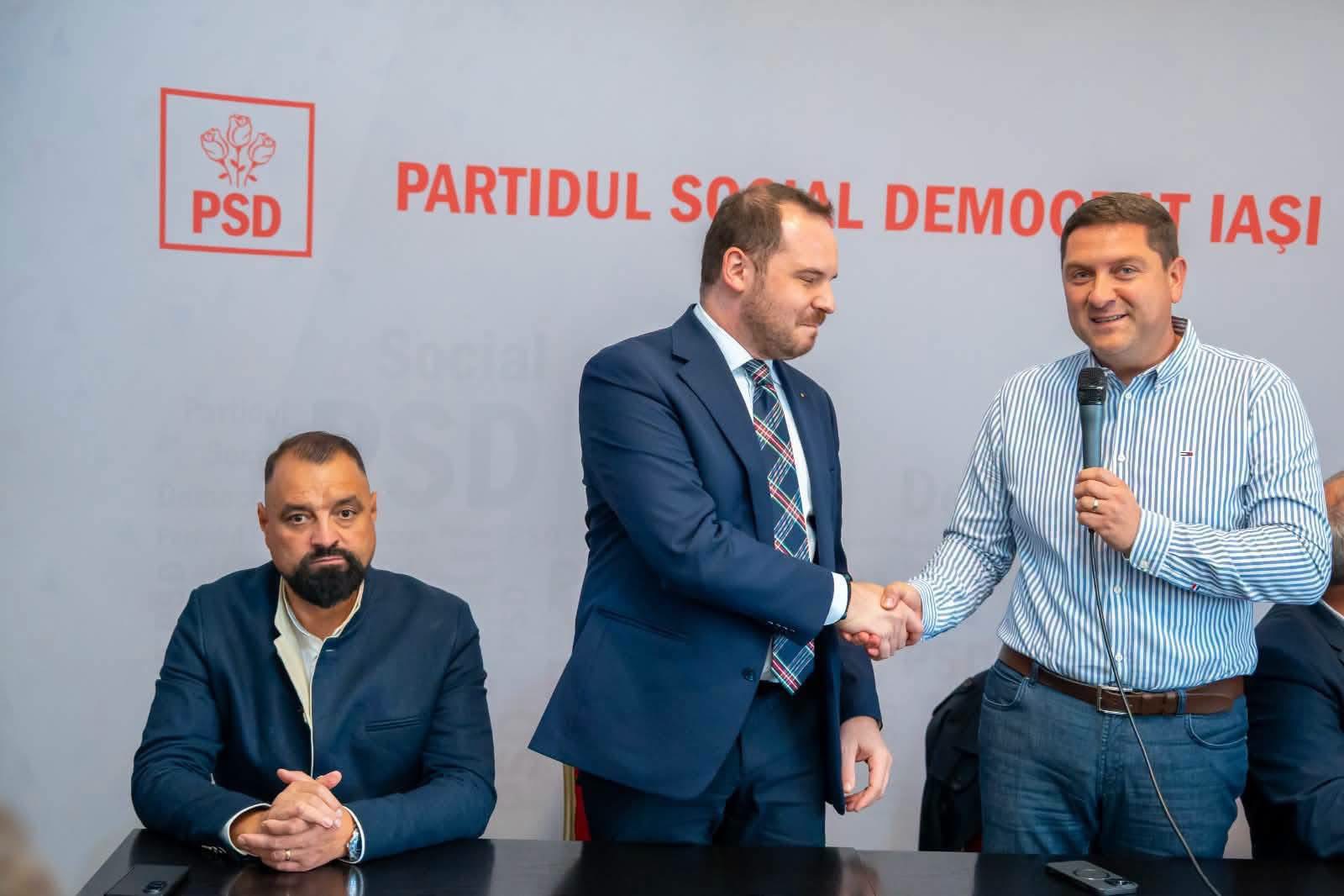 Doi bărbați își dau mâna în fața siglei PSD, iar un al treilea stă așezat la masă