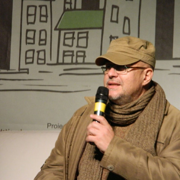 Poetul Radu Andriescu vorbind la microfon, cu șapcă și fular