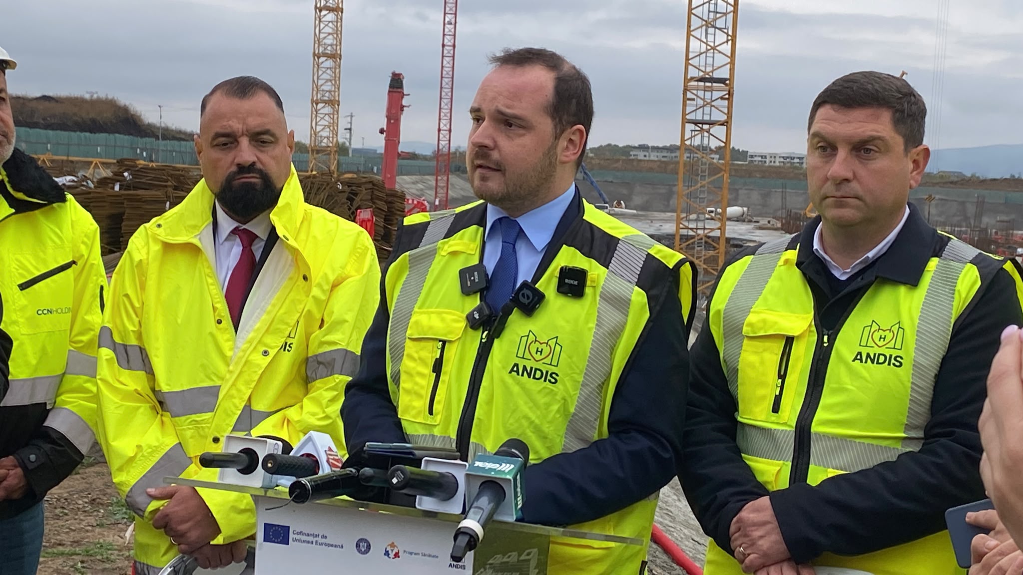 Conferință de presă pe un șantier, cu mai mulți bărbați în veste reflectorizante